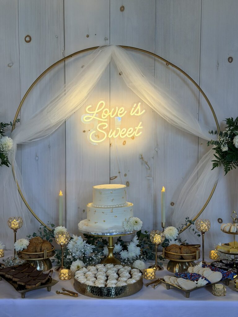 Two-tier wedding cake with gold leaf beneath a glowing Love is Sweet neon sign, framed by a gold hoop with tulle and white florals