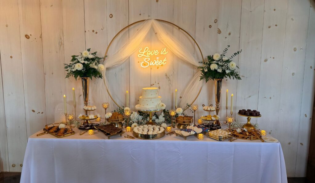 Styled wedding dessert table with gold hoop backdrop, tulle, white florals and a glowing Love is Sweet neon sign
