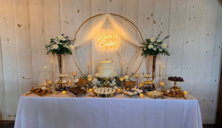 Styled wedding dessert table with gold hoop backdrop, tulle, white florals and a glowing Love is Sweet neon sign