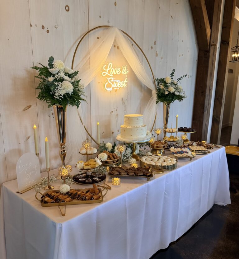 Side view of a styled wedding dessert table with gold vases, candlesticks, a Love is Sweet welcome sign and cannoli in a rustic barn venue